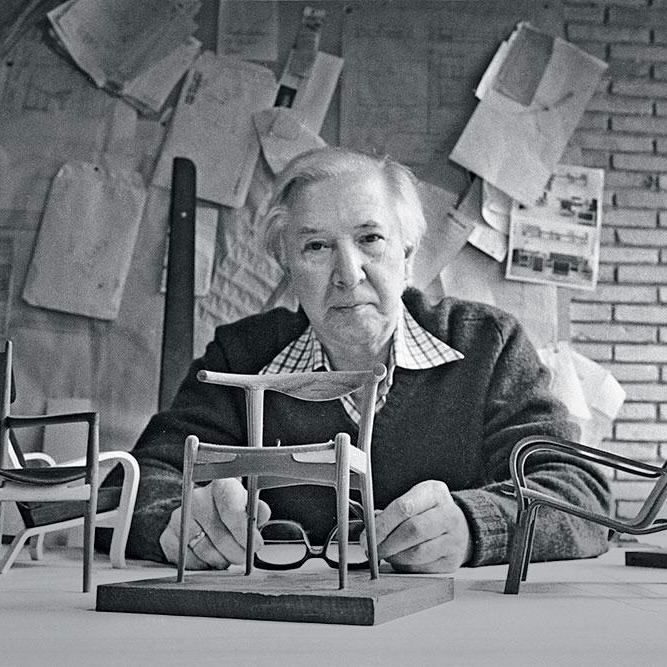 Чёрно‑белая сцена: человек за столом с миниатюрами кресел и развешанными на стене эскизами и заметками дизайна.