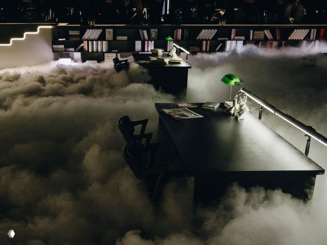 Крупный план рабочего стола в инсталляции: тёмная поверхность, книги, зелёная настольная лампа и густой туман, создающий сюрреалистическую сцену.