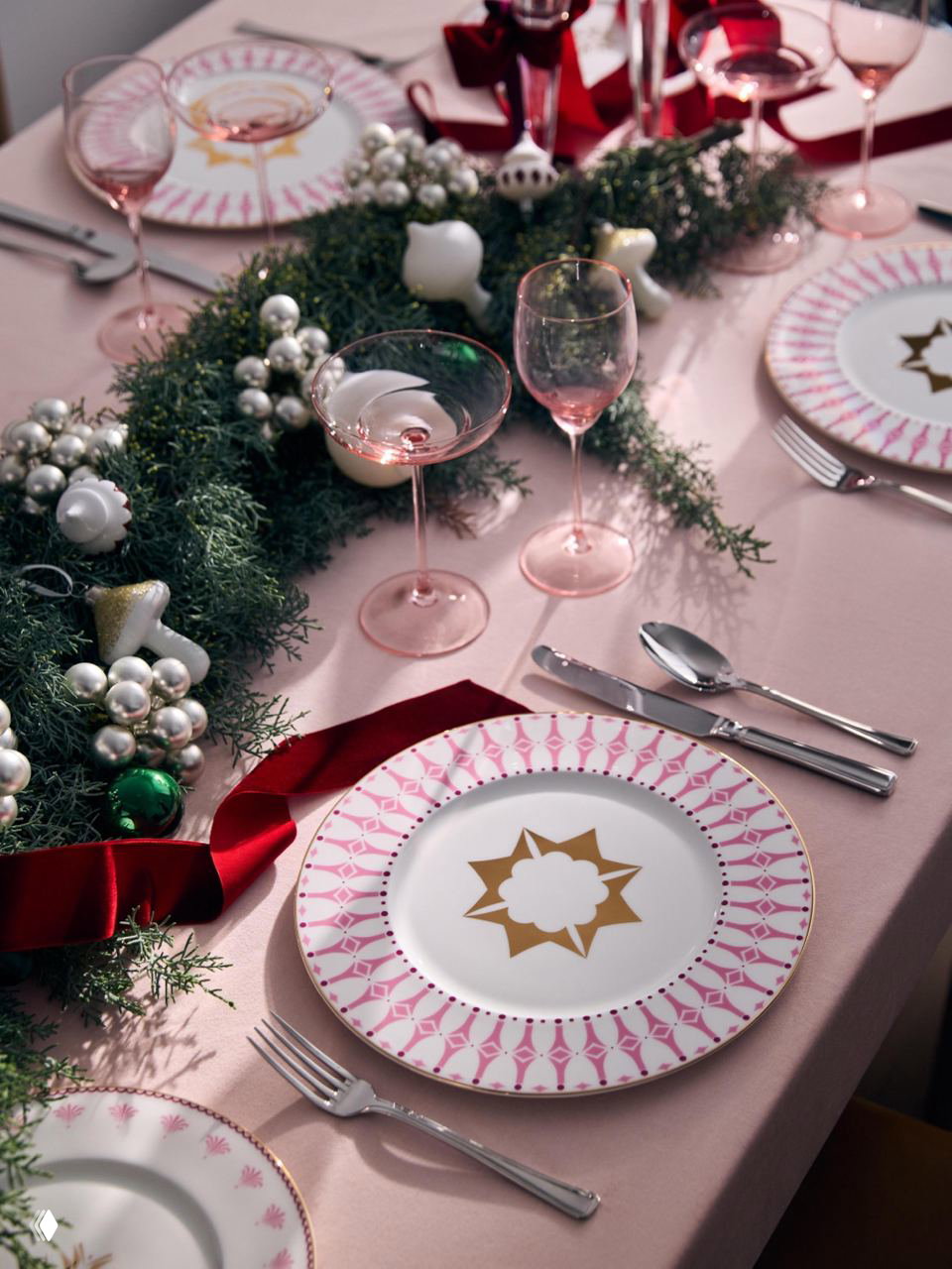 Романтичная праздничная сервировка с тарелками из коллекции «Пурпур»: розовые и золотые акценты, рождественская гирлянда и стеклянные бокалы на розовой скатерти.