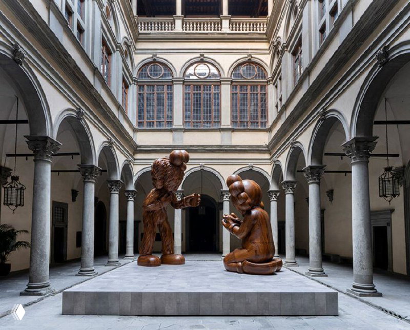 Крупный план композиции «Послание» во дворе: стоящий персонаж Companion и сидящая BFF из дерева на фоне архитектуры палаццо Строцци.