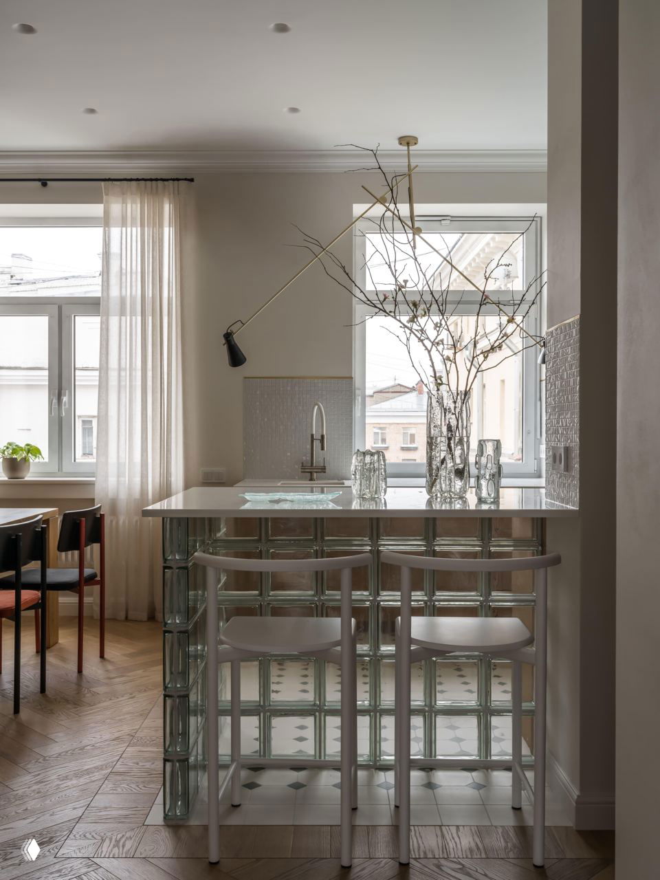 Кухонная барная зона у окна: стеклоблоки под столешницей, белая столешница, стеклянные вазы и большое окно, обеспечивающее мягкое естественное освещение.