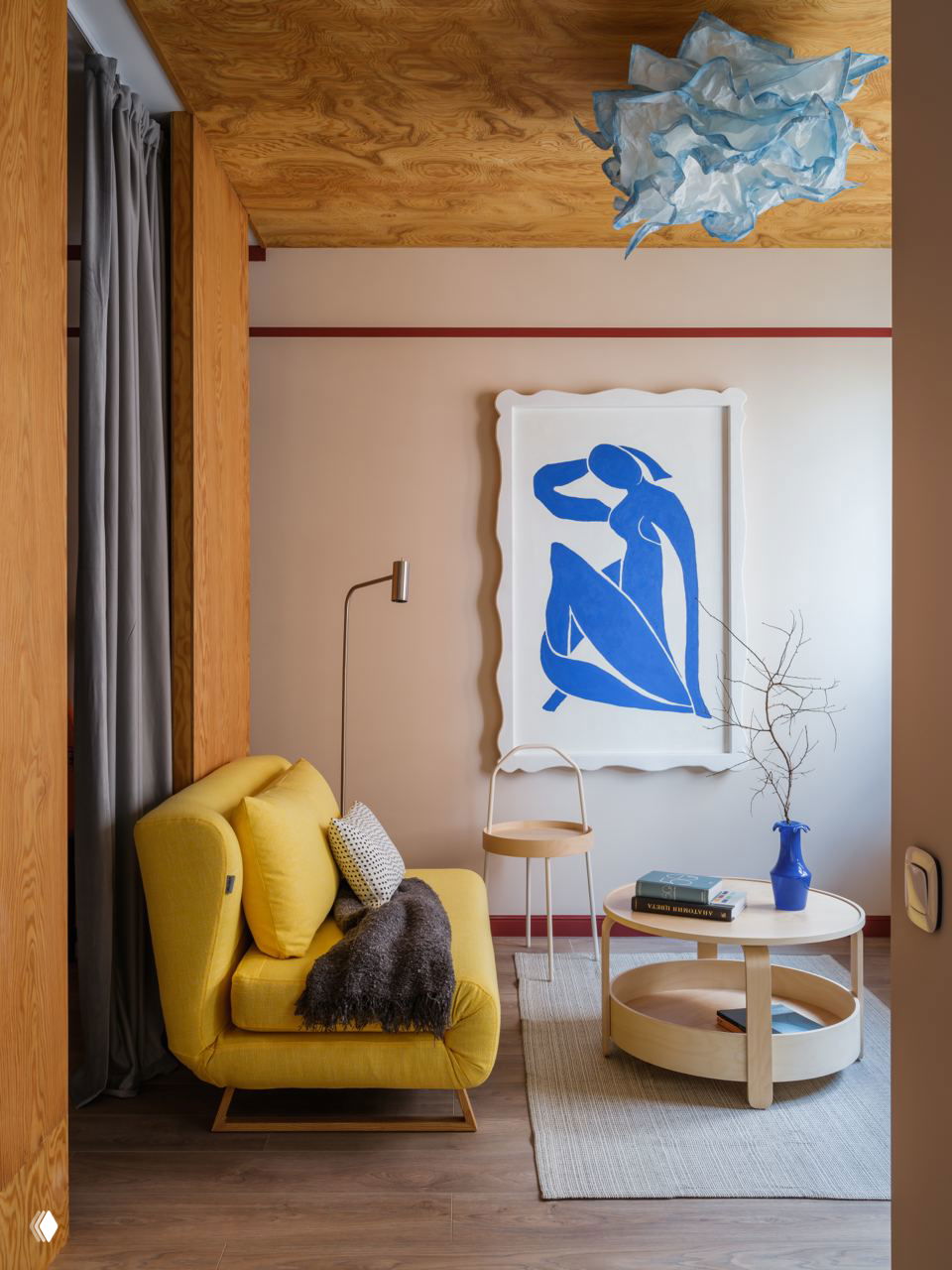 Небольшая комната с жёлтым креслом, круглым журнальным столиком и крупной настенной иллюстрацией в синем, тёплые и яркие пятна в нейтральном пространстве.