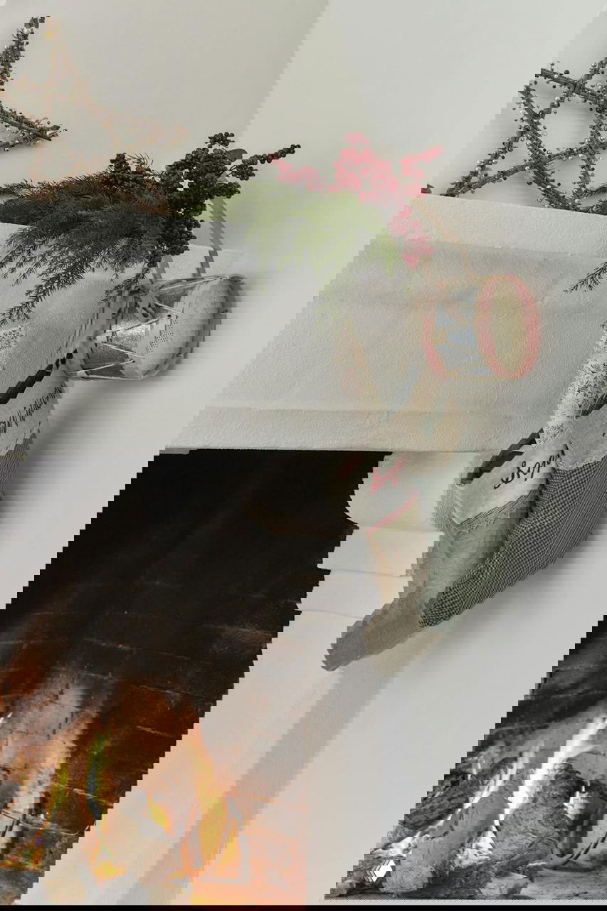 Деталь каминной полки: рождественские носки, зелёная ветка и декоративный барабан на белом фоне рядом с горящим огнём в камине.