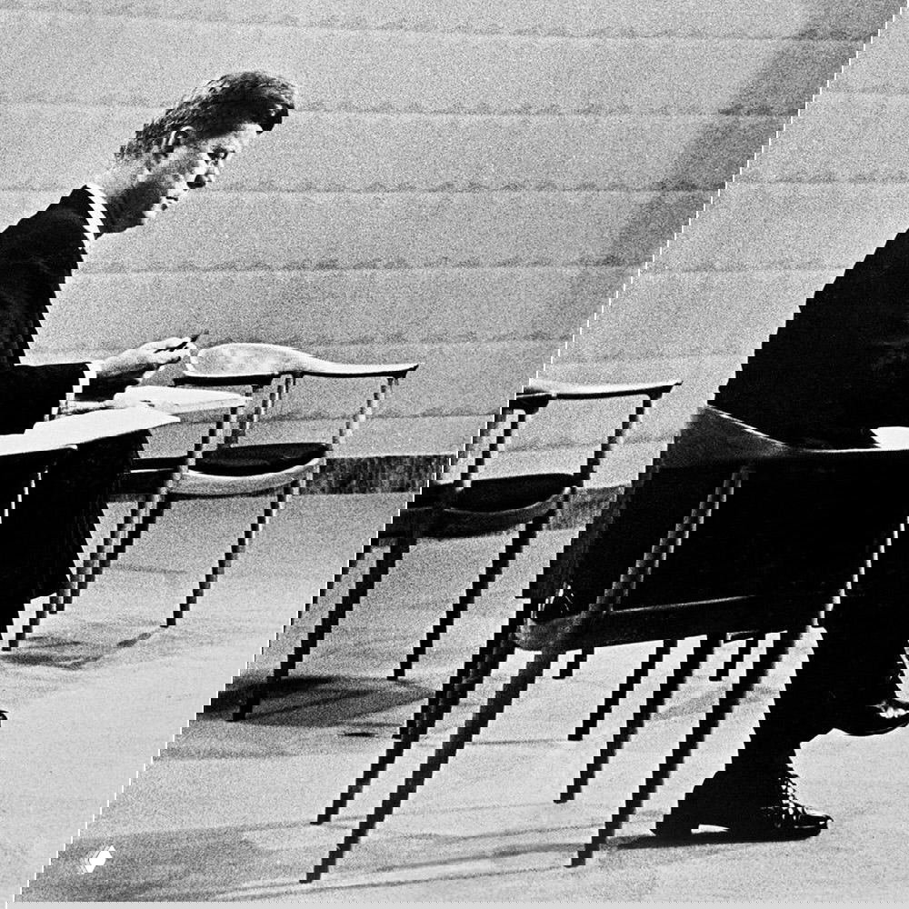 Чёрно‑белая телевизионная сцена 1960-х: мужчина в студии, сидящий в элегантном деревянном кресле, рядом другие схожие модели.