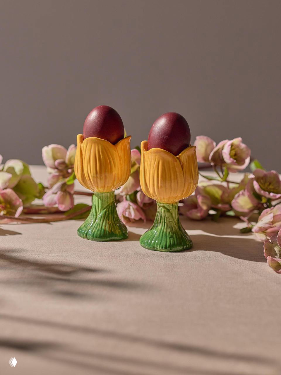 Две тюльпанообразные подставки для яиц из коллекции Blossom с окрашенными пасхальными яйцами на фоне пастельных весенних цветов и мягкого светового осветления.