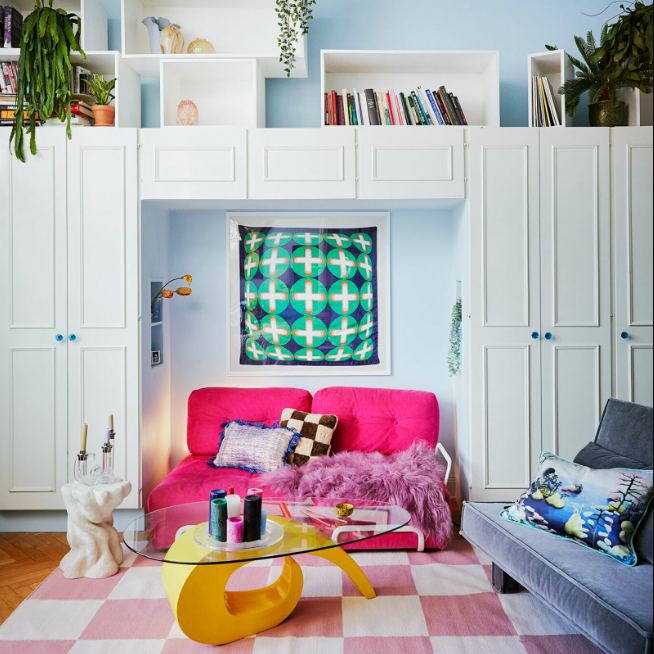 Светло-розовый компактный диван с пушистой подушкой и мягкой игрушкой на фоне нейтральной стены и минималистичной настенной лампы, домашняя атмосфера.