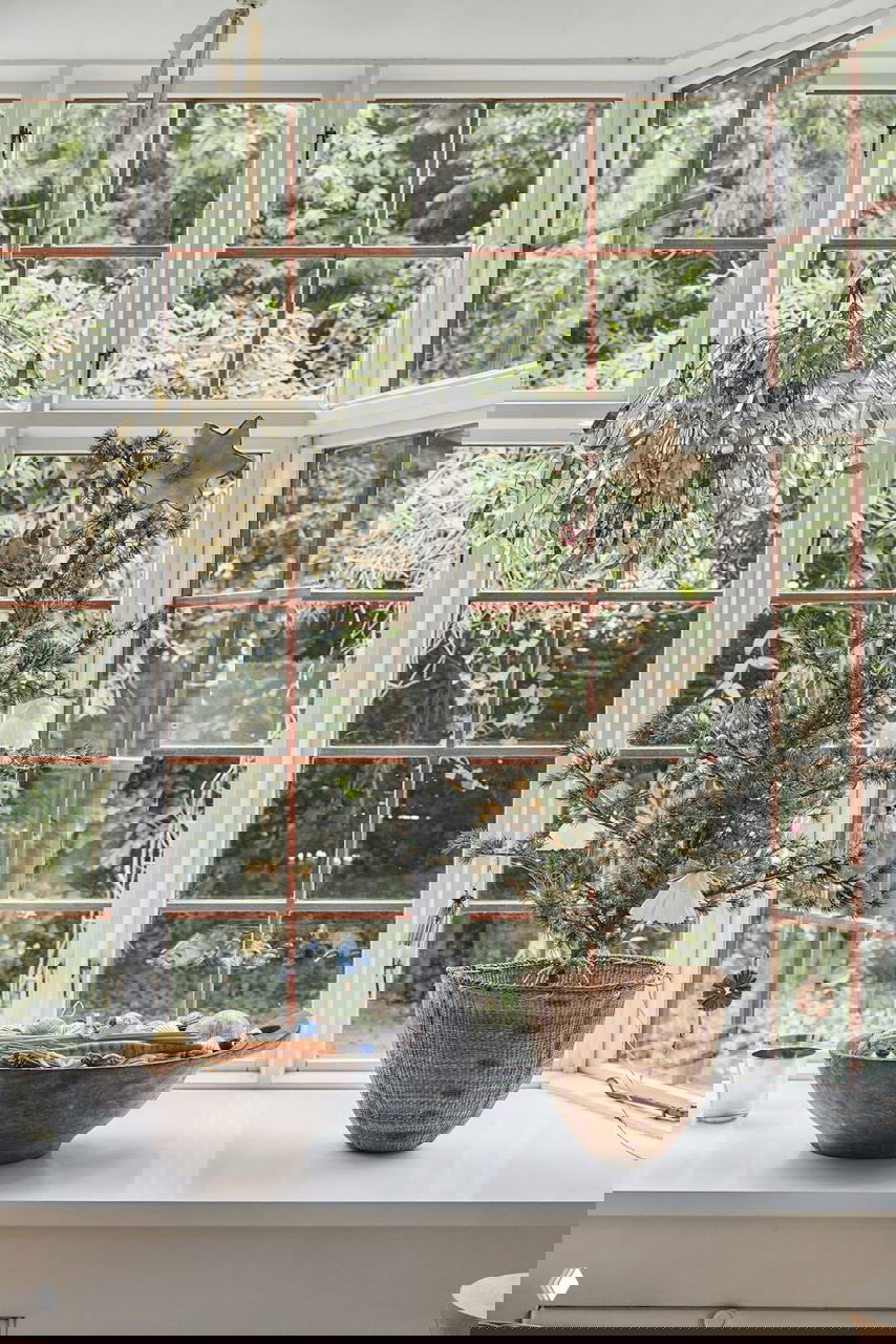 Подоконник с мини-ёлкой в плетёной корзине, миска с украшениями и большая панорамная рама окна с видом на зелёный сад за домом.