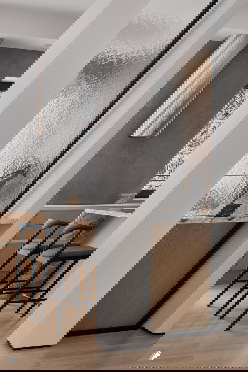 Кухонная зона с барными стульями, мраморной столешницей и матовым текстурным стеклом-перегородкой; светлое деревянное напольное покрытие и лаконичные формы.