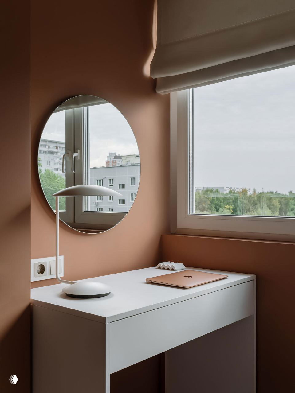 Рабочий уголок у окна с круглым зеркалом и столом на фоне стен в тёплом коричнево-терракотовом оттенке, мягкий дневной свет сквозь окно.