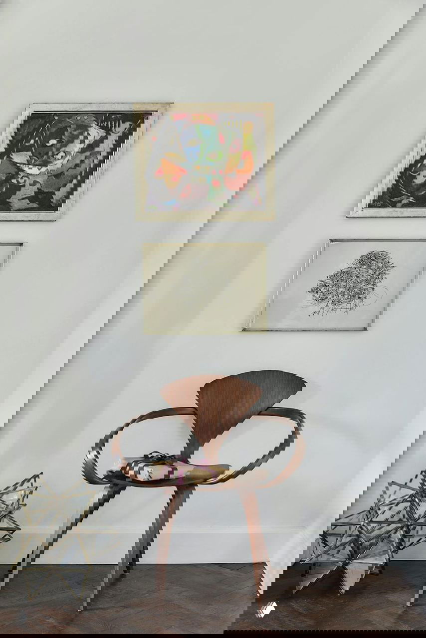 Деревянный дизайнерский стул у белой стены с картинами и декоративной фигурой рядом, аккуратный интерьерный уголок в светлых тонах.