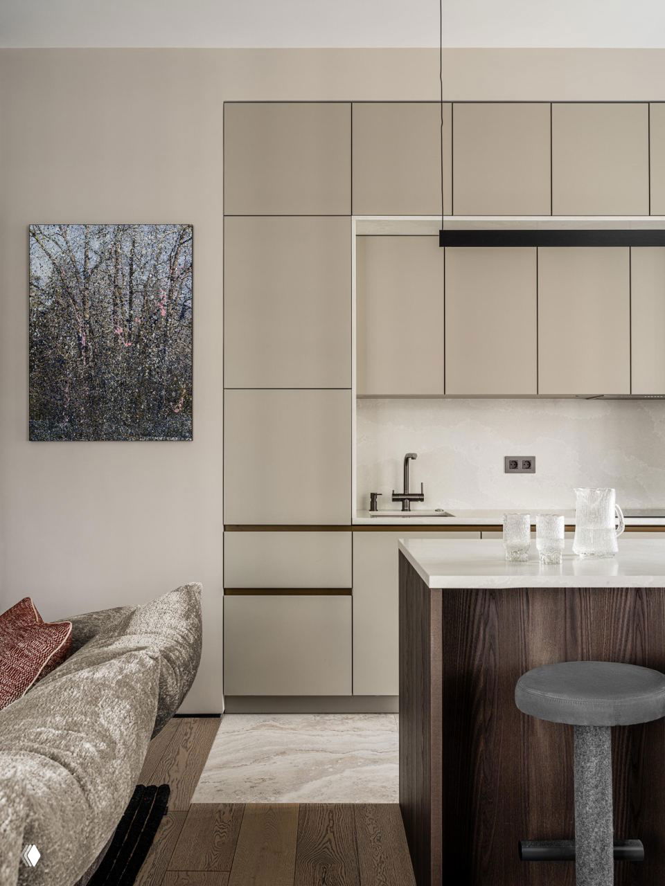 Кухонный фрагмент со встроенными фасадами в приглушённом бежевом, остров и мягкий диван в зоне гостиной рядом с ковром.