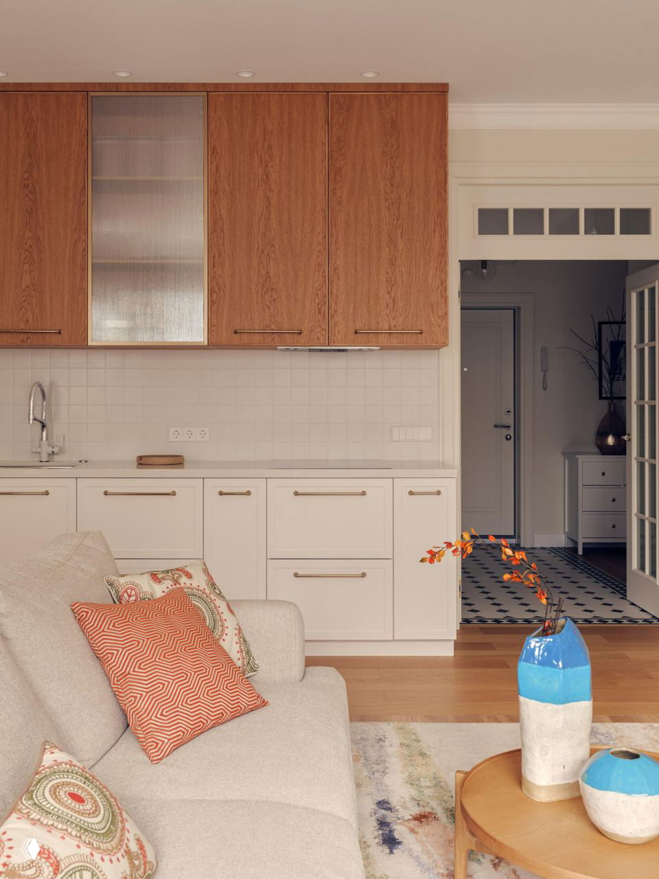 Кухонная зона, белые нижние шкафы и верхние фасады из натурального дерева, акценты терракоты в текстиле и керамике, единый тёплый интерьерный стиль.