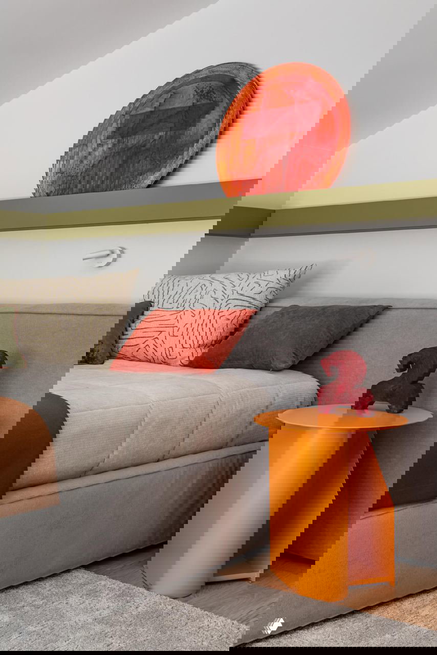 Спальня с мягким серым изголовьем, яркими акцентами в оранжевом и терракотовом цвете: небольшой круглый оранжевый столик и декоративная пластика на стене.