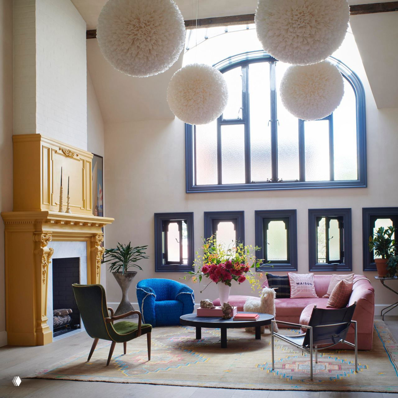 Роскошный бархатный розовый диван у большого арочного окна и золотистого камина; в комнате кресла, журнальный стол и яркая флористика создают интерьер мечты.