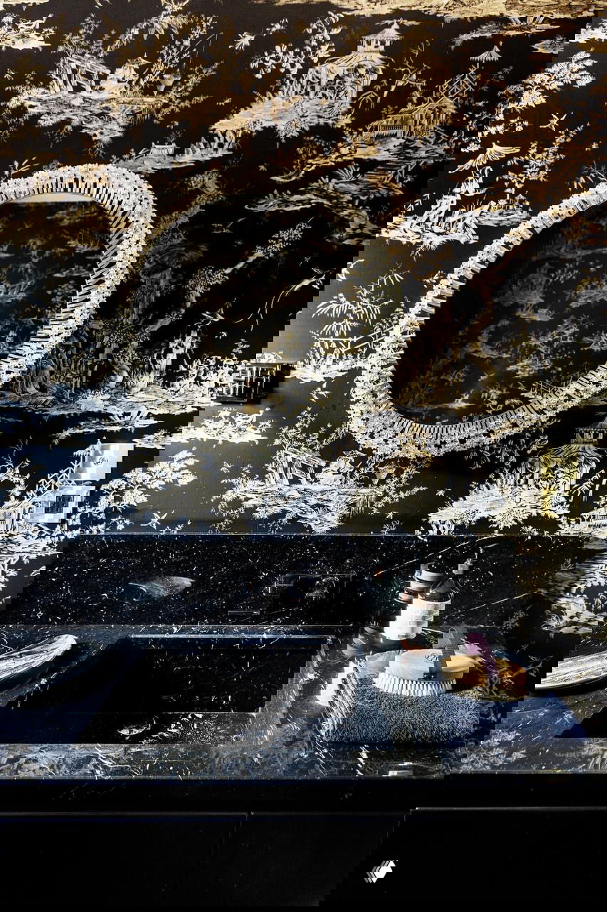 Чёрные обои с золотыми тонами и традиционными сюжетами, круглое зеркало с рельефной рамой и тёмная мраморная раковина на столешнице.