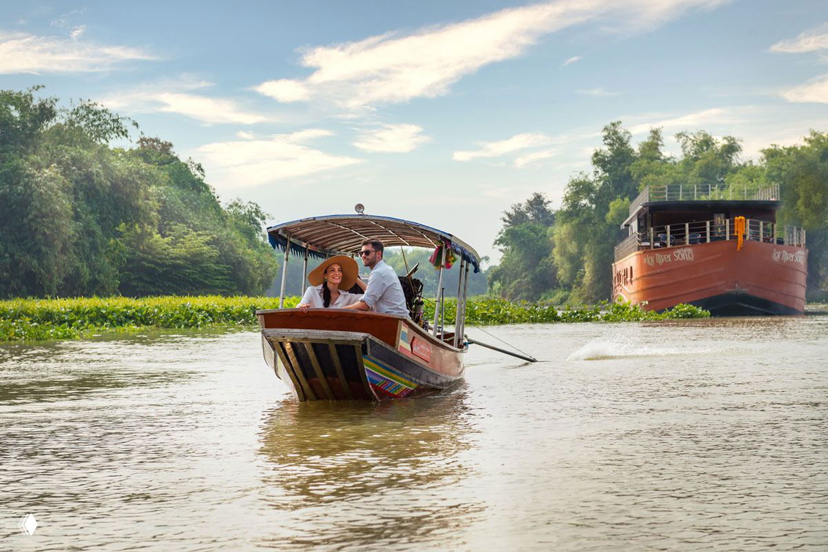 Традиционная тайская моторная лодка на реке Чхаопрайя с парой туристов на носу, вокруг плавучая растительность и баржа вдалеке на реке.