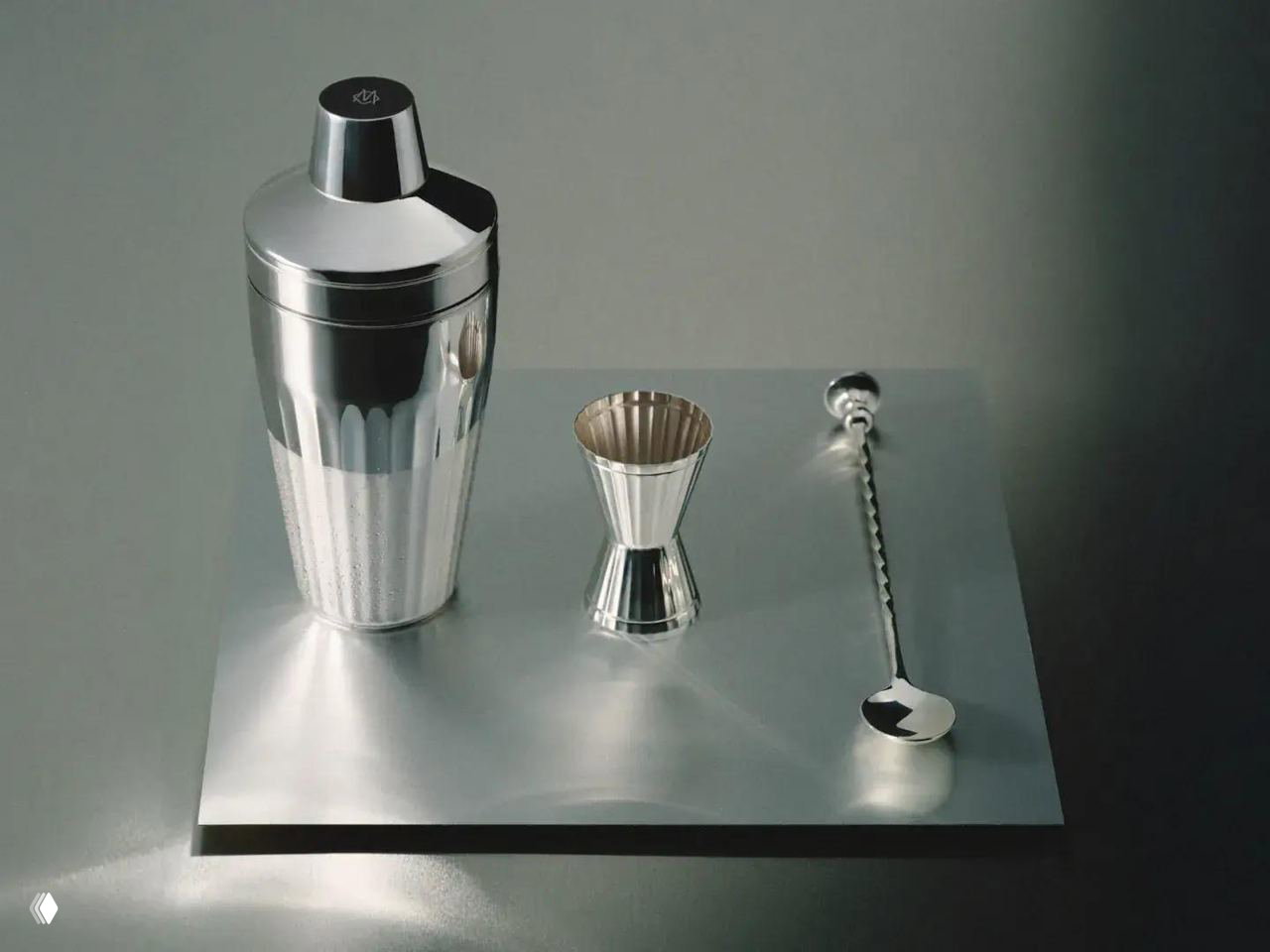 Посеребренный барный шейкер, джиггер и скрученная барная ложка на нейтральном фоне, демонстрирующие зеркальную отделку и текстуру металла.