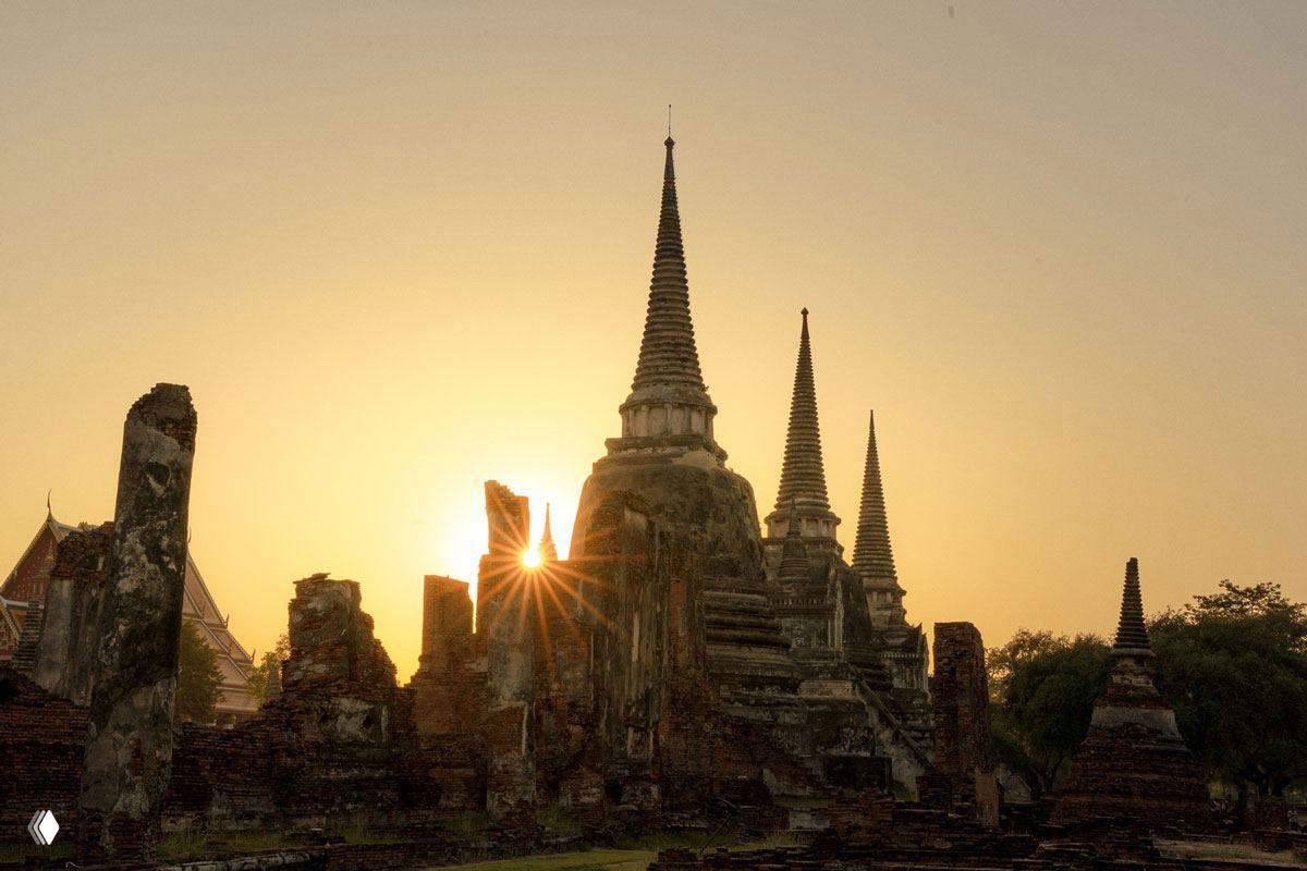 Руины древней Аюттайи при закате: силуэты чеди и ступенчатых шпилей на фоне тёплого неба и заходящего солнца над историческим комплексом.