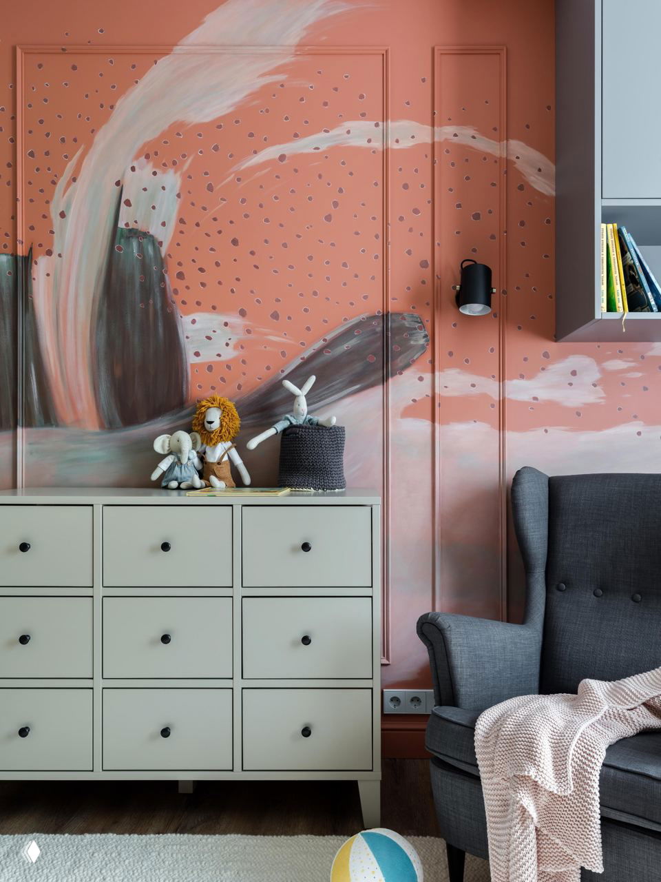 Детская или гостиная с акцентной рукотворной росписью на стене в тёплых коралловых тонах, плетёные игрушки и светлая мебель на переднем плане.