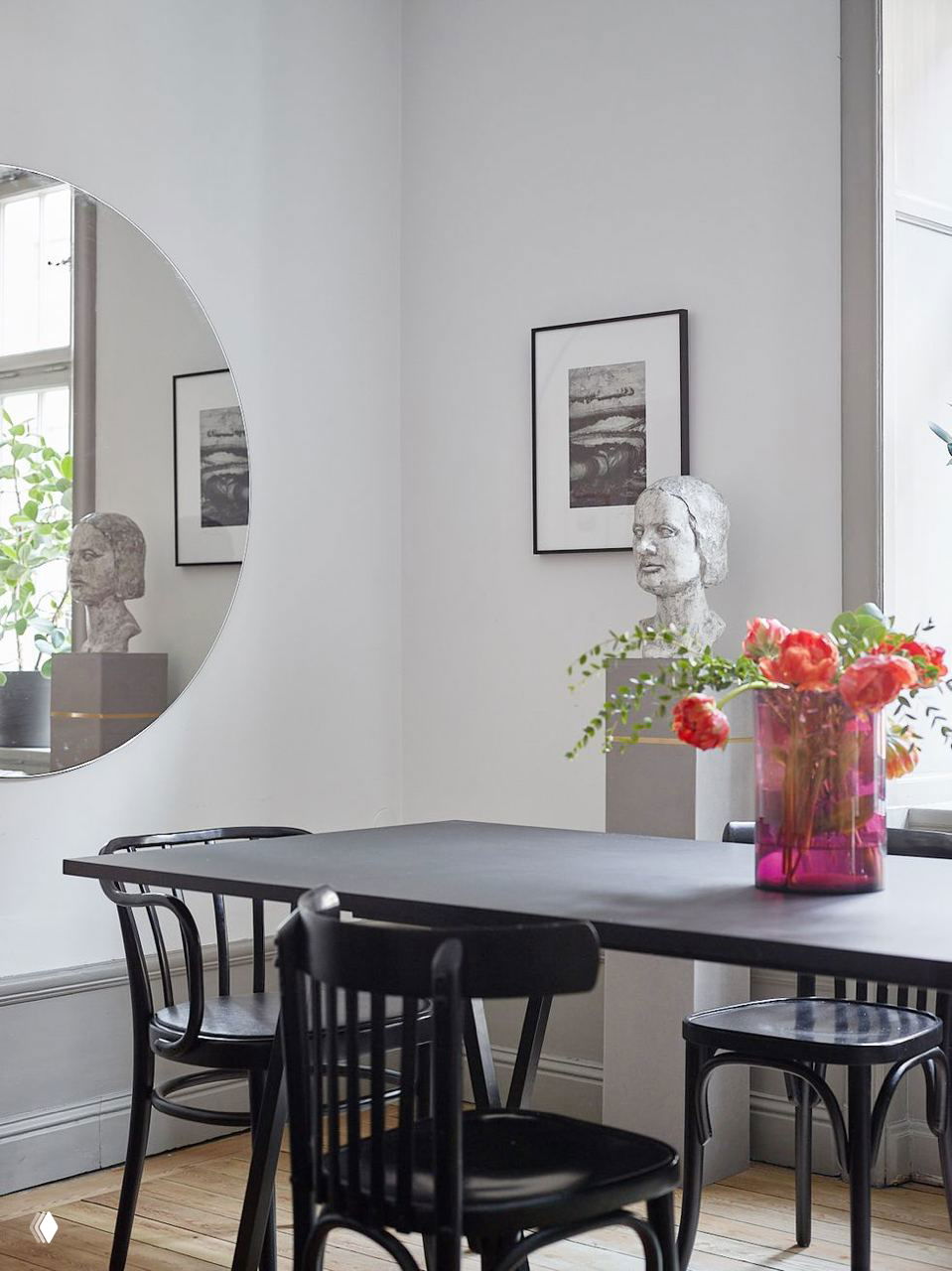 Обеденная зона с темным столом и несколькими венскими стульями на фоне круглого зеркала, скульптуры и цветочной композиции.