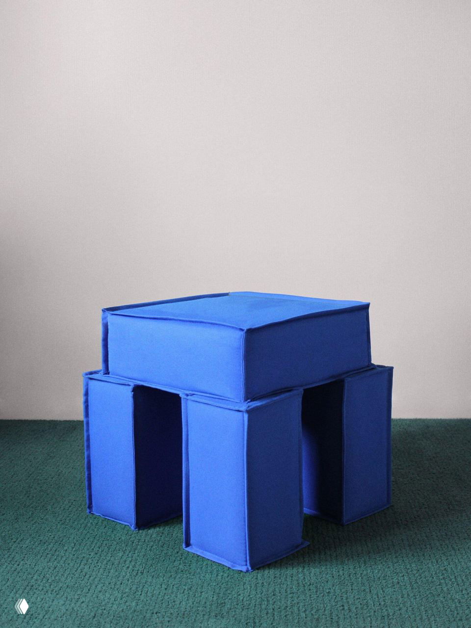 Насыщенно‑синяя модульная табуретка с мягкой обивкой на зелёном ковровом покрытии, строгие геометрические формы и минималистичный дизайн в интерьере.