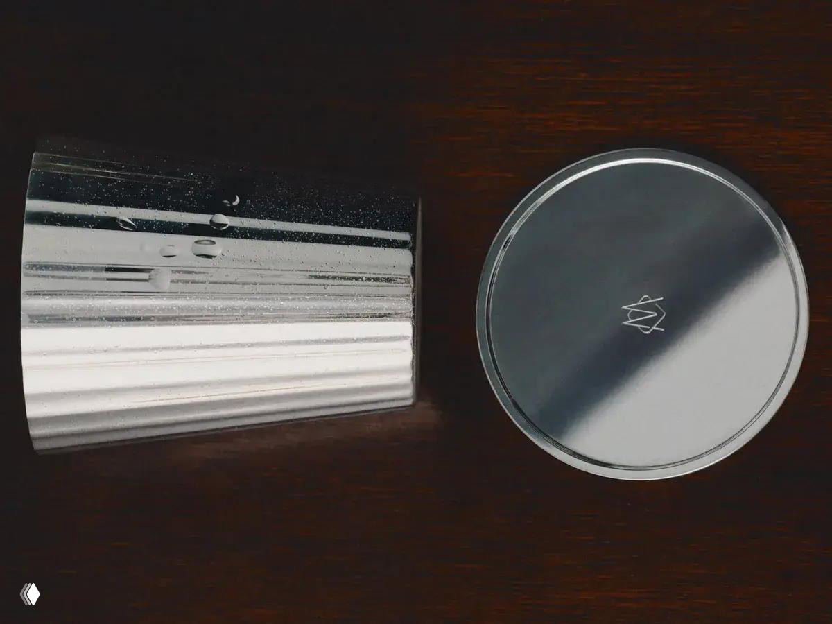 Детали посеребренного стакана и круглой крышки с выгравированным логотипом на темном фоне; видны рифлёные поверхности и отражения металла.