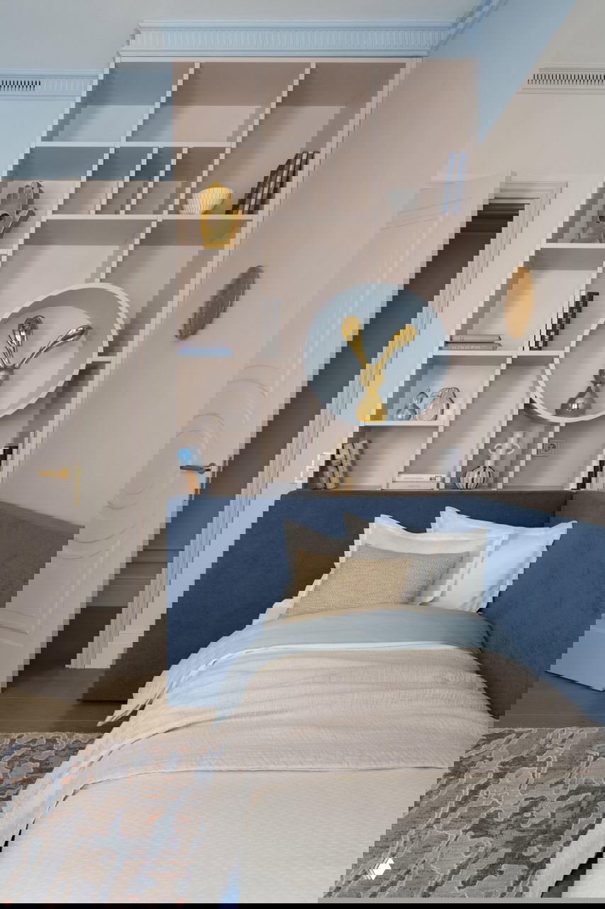 Детская/гостевая зона с синим мягким изголовьем, открытыми полками и декоративными объектами — аккуратные акценты в приватной части квартиры.