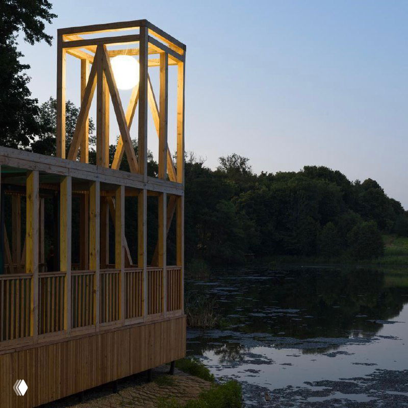 Деревянный прибрежный павильон на опорах у озера в сумерках, подсвеченный изнутри, с отражением воды и линией леса на заднем плане.