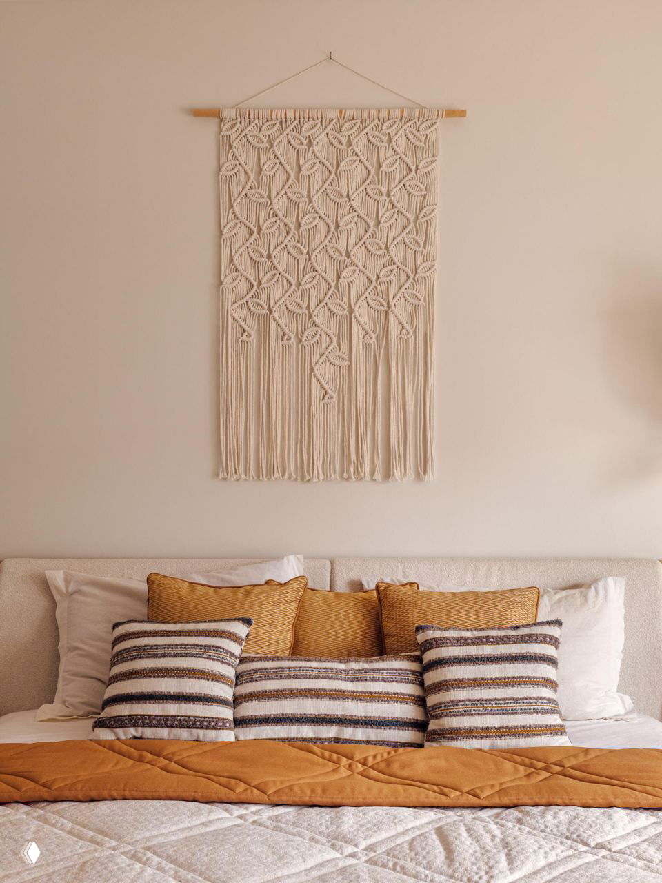 Кровать с декоративными подушками в полоску и терракотовых тонах, макраме-панно над изголовьем и мягкое постельное покрытие теплого оттенка.