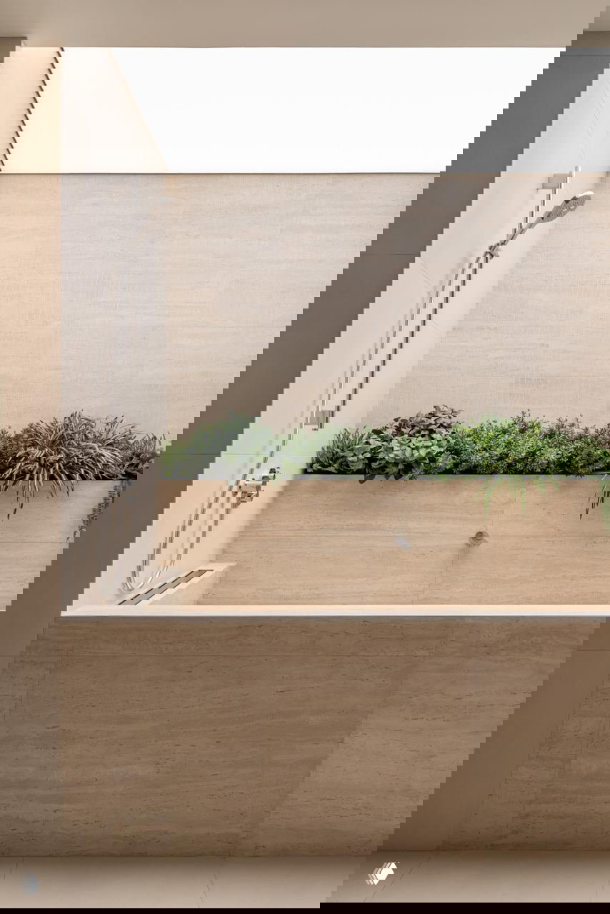 Санузел с большой ванной в отделке натуральным камнем и встроенной зелёной клумбой, мягкое равномерное верхнее освещение и минималистичные фитинги.