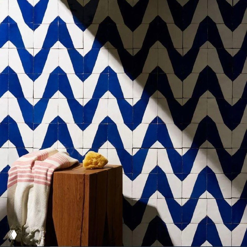 Синяя и белая плитка в узоре «шеврон», рядом деревянная табуретка с полотенцем и губкой, солнечный свет подчёркивает фактуру керамики