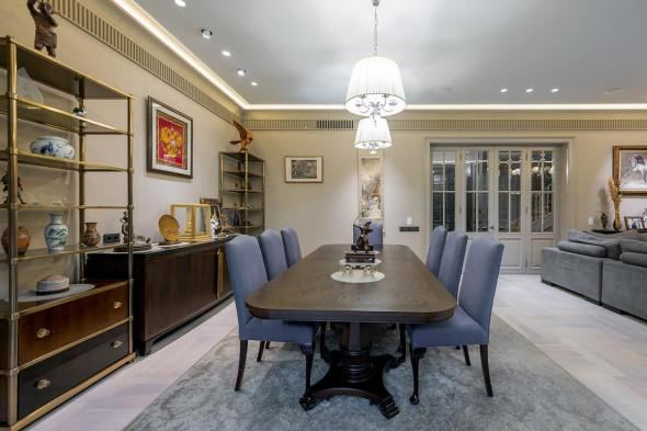 Столовая зона с большим деревянным столом, стульями и стеллажами с декоративными предметами, оформление в тёплых тонах и текстиле.