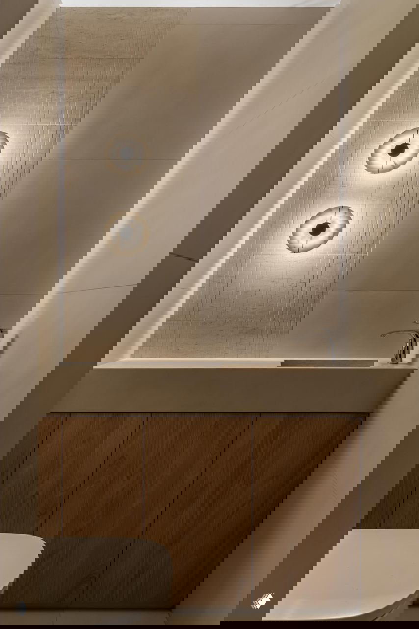 Нейтральный санузел со скрытой подсветкой, деревянным шкафчиком и круглой настенной подсветкой — минималистичный и спокойный интерьерный приём.