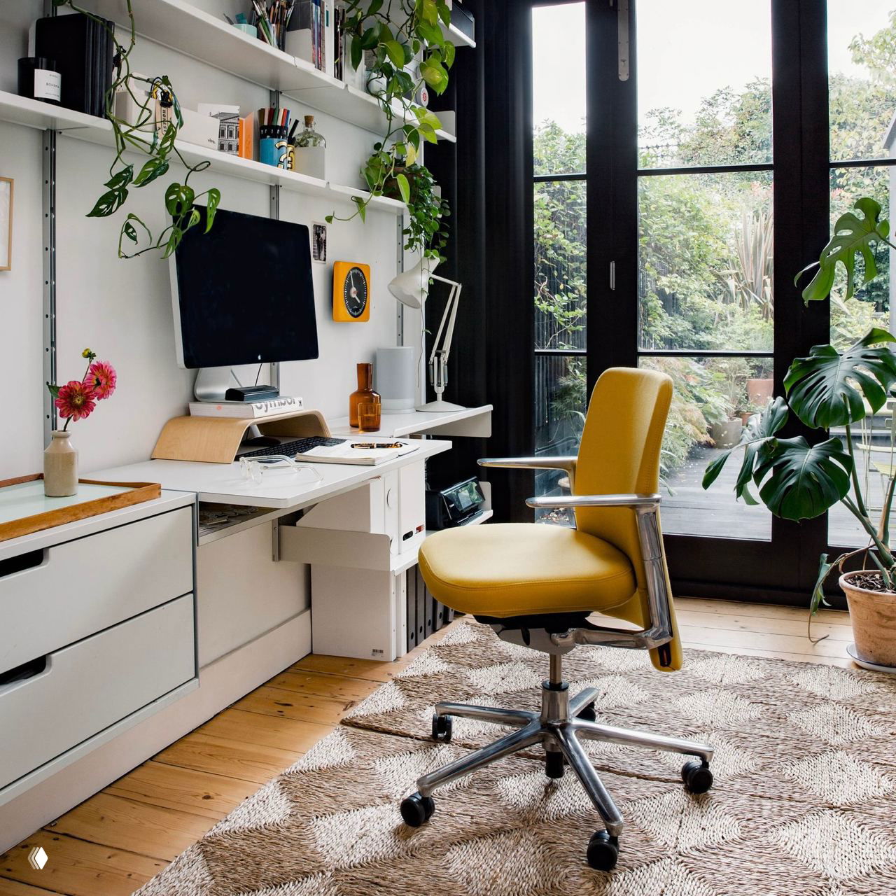 Уютный рабочий уголок с жёлтым креслом на колёсиках, белый стол и подвесные растения у больших чёрных дверей-окон.