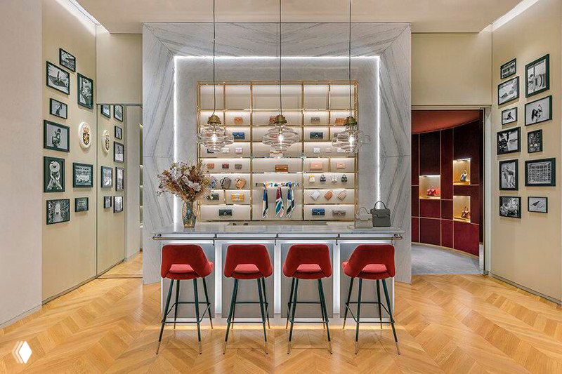 Интерьер бутика Delvaux с центральной зоной отдыха и яркими сиденьями: элегантные полки с сумками, деревянный паркет и мягкая подсветка витрин.