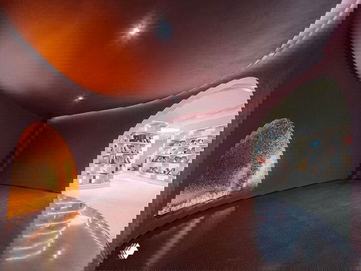 Тёмный куполообразный зал с арочным входом и золотистой нишей‑печью, переходящий в светлую зону с книжными полками — отсылка к этапу обжига.