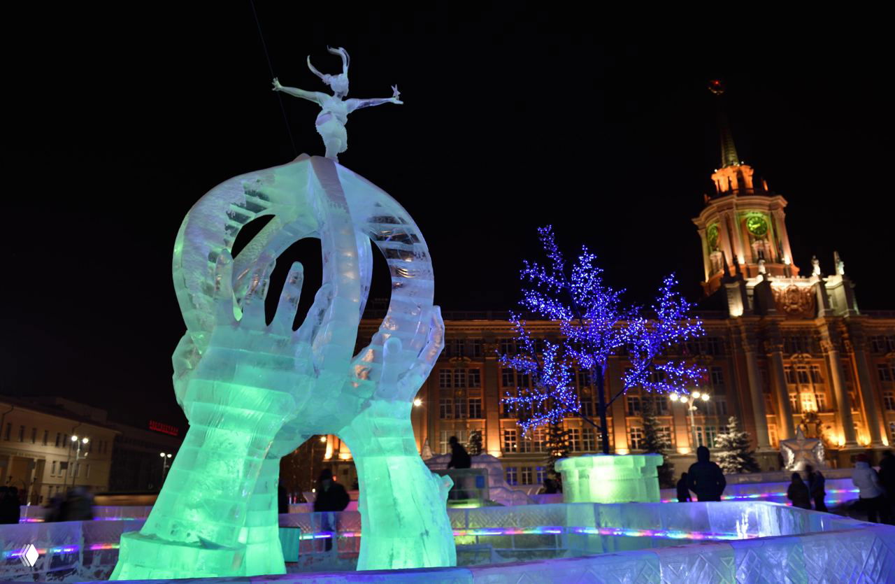 Подсвеченная ледяная скульптура на городском празднике вечером, с синими огнями на деревьях и историческим зданием на заднем плане, прохожие вокруг