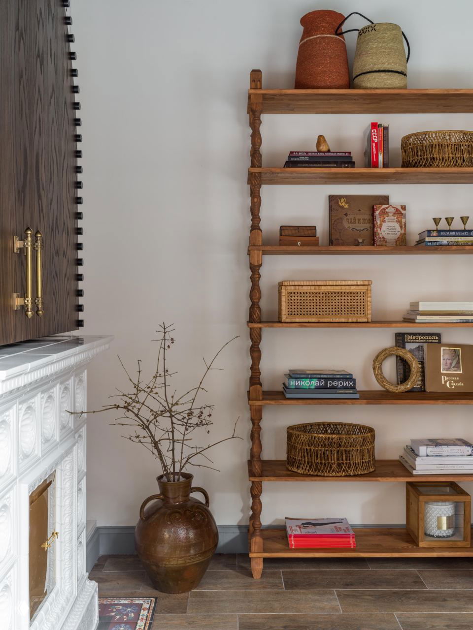 Деревянный стеллаж с плетёными корзинами и книгами, рядом большая керамическая ваза с сухими ветками и часть белой резной облицовки камина.