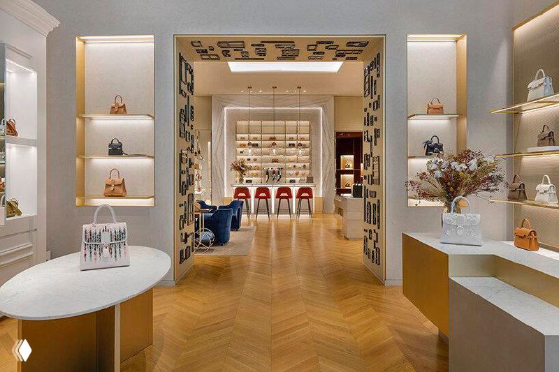 Барная стойка бутика Delvaux с мраморной столешницей и бледно‑золотыми деталями на фоне полок с аксессуарами и декоративными композициями.