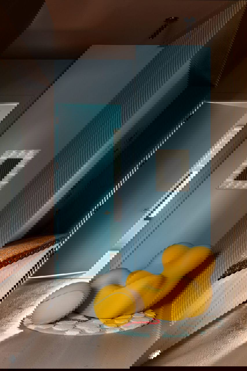 Прихожая с голубой дверью и тёплым коричневым потолком, скульптурное жёлтое кресло и круглый коврик — неожиданные формы и яркие акценты в проходной зоне.