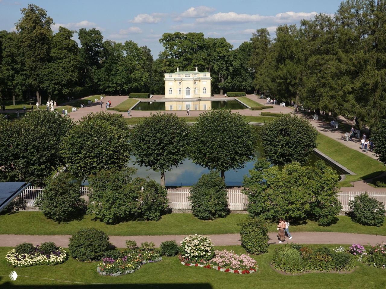 Парк Царского Села с симметричным прудом и небольшим павильоном, аккуратные аллеи, цветники и прогуливающиеся люди летом.