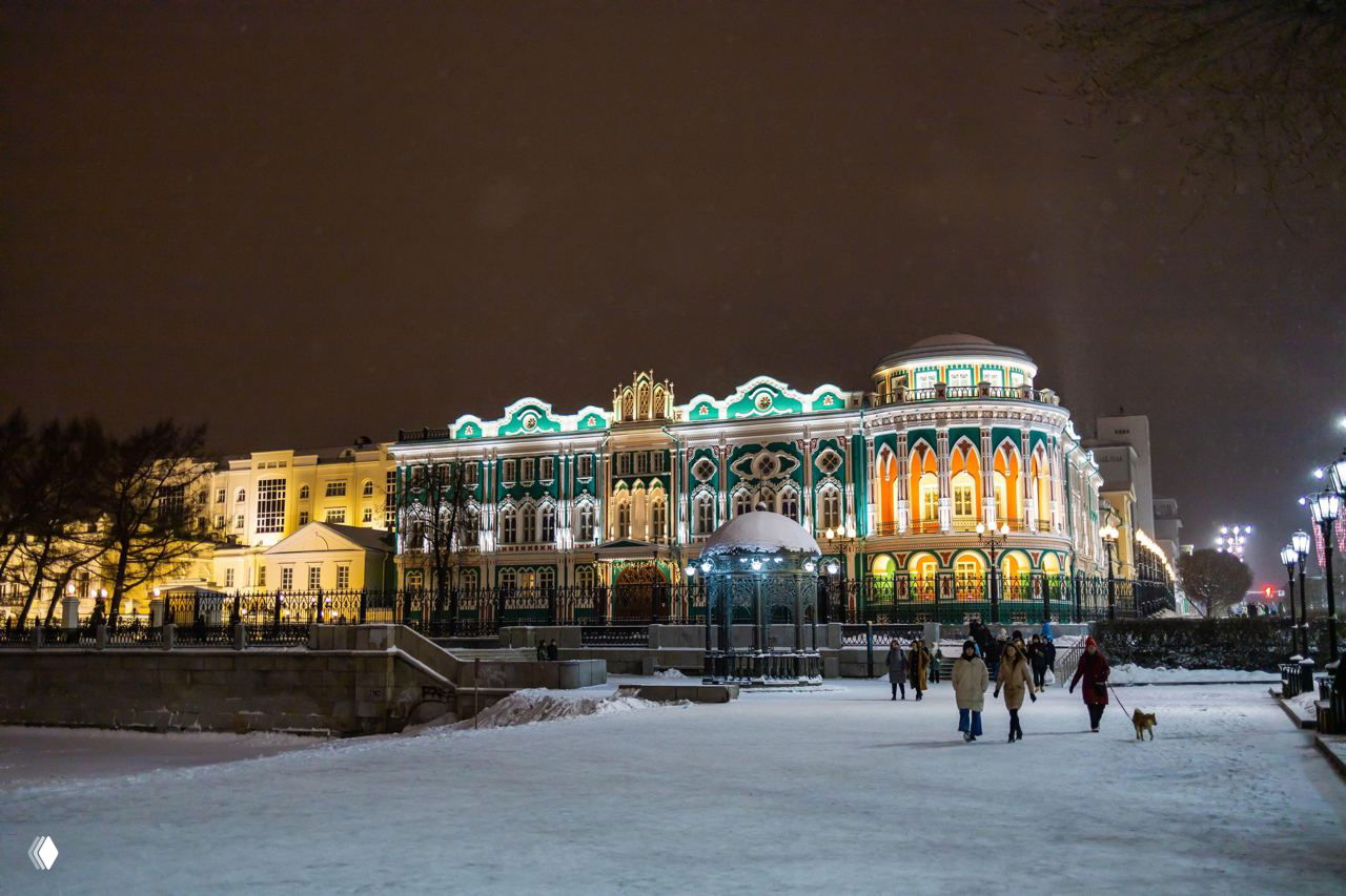 Дом Севастьянова вечером, фасад здания подсвечен, рядом заснеженная набережная и гуляющие люди — стильная архитектура города зимой
