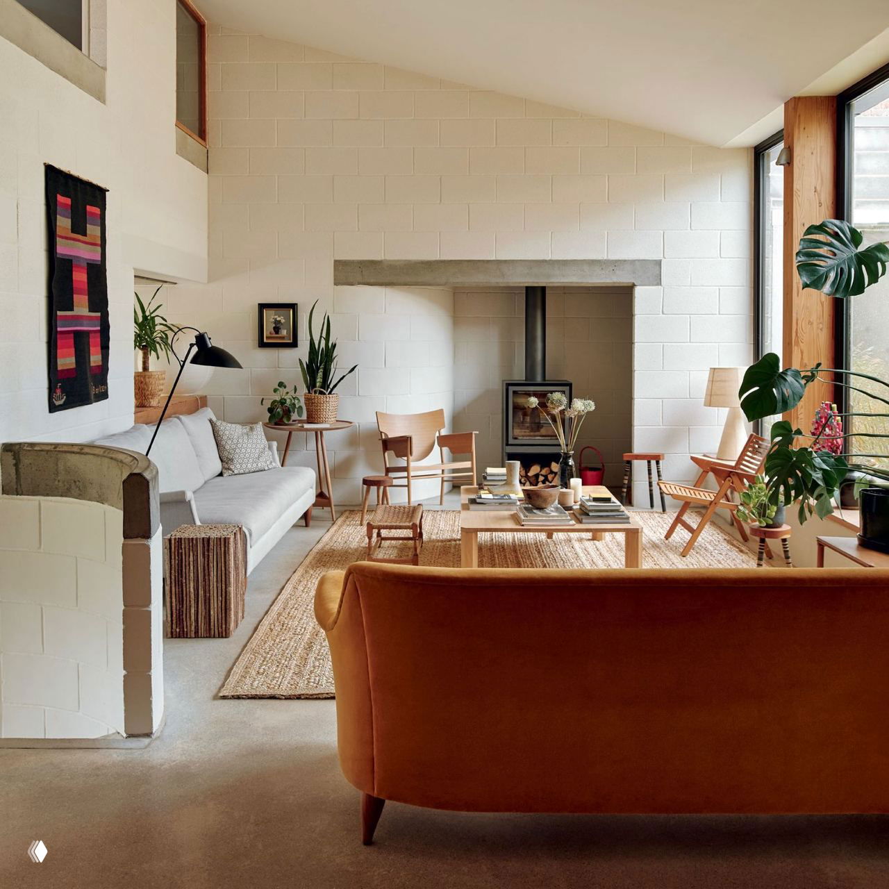 Гостиная с камином, диванами и плетёным ковром; комнатные растения на подставках и в горшках создают зелёный акцент и тёплый интерьер.