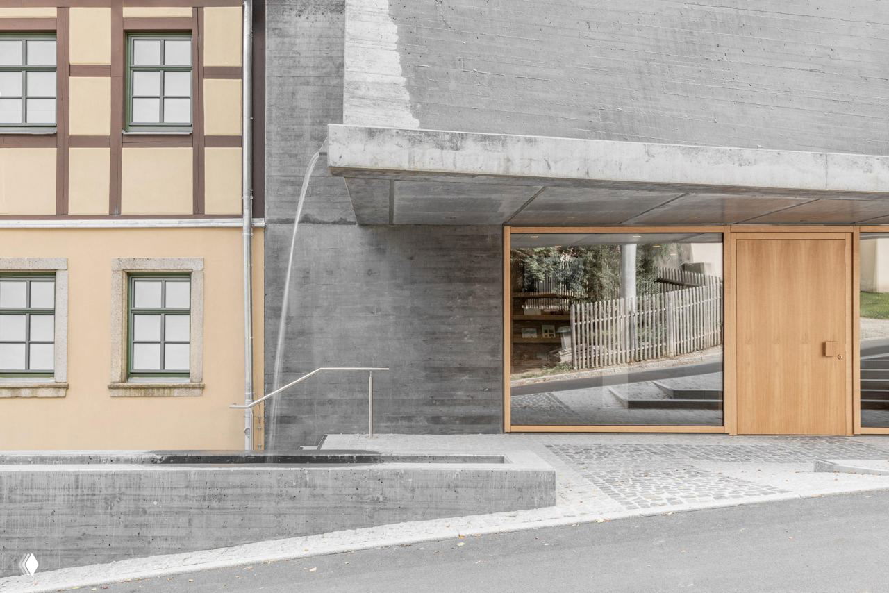 Деталь фасада: желоб для дождевой воды стекает по бетонной поверхности в небольшой бассейн у входа, рядом витрина и деревянная дверь