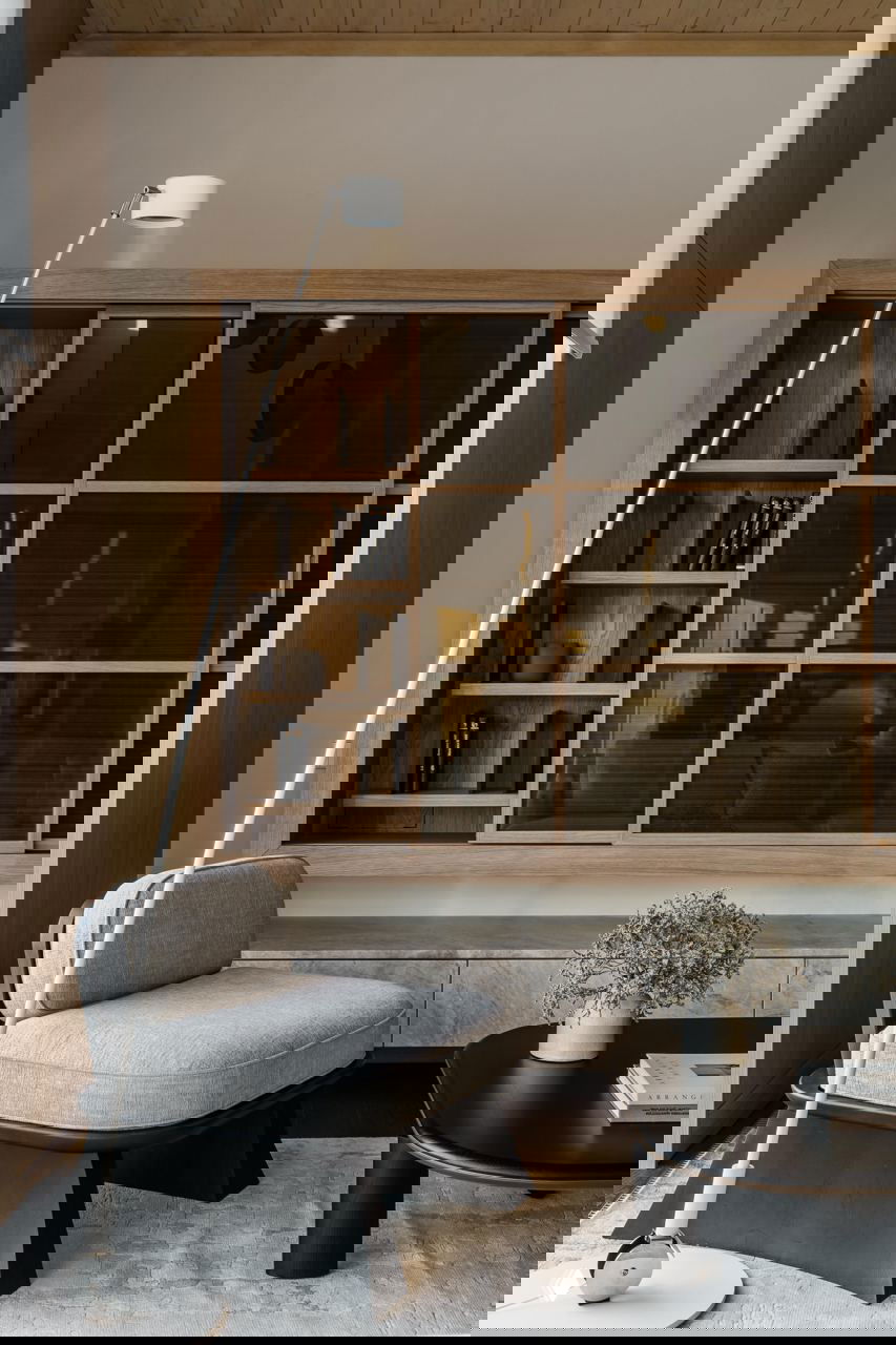 Читальная зона с низким креслом и закрытой стеклом деревянной библиотекой за ним, спокойные тона и мелкие предметы декора на столике.