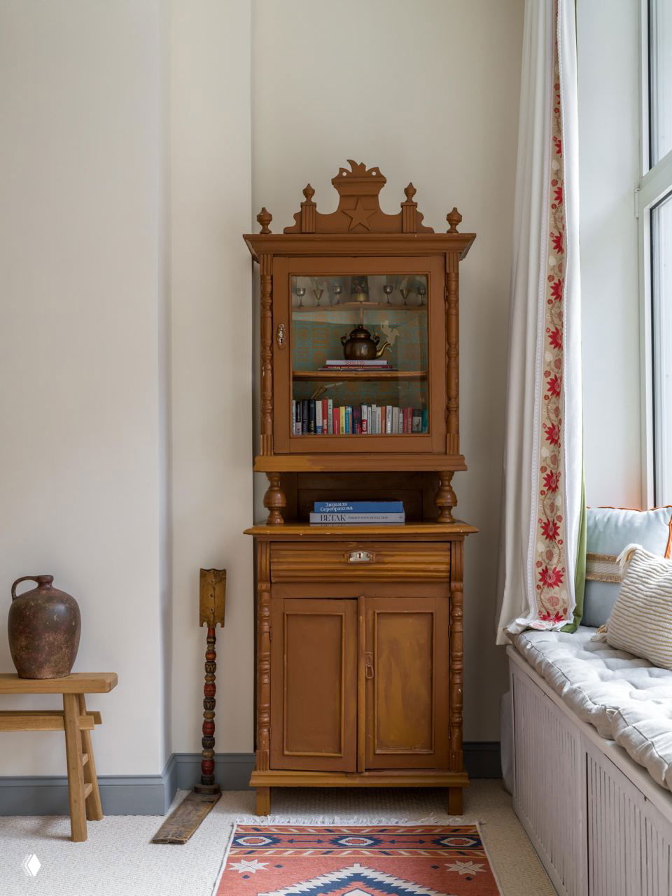Высокий деревянный шкаф‑витрина у окна, рядом с подоконником с мягким сиденьем и текстильным рисунком на коврике в традиционном стиле.