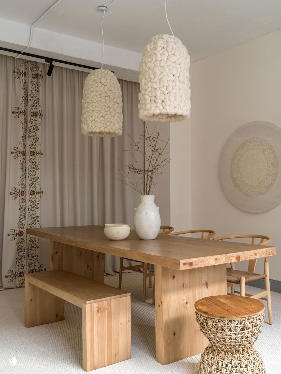 Большой деревянный стол и скамьи в натуральной отделке под мягкими подвесными светильниками с крупной текстурой и вазой с ветками в центре.