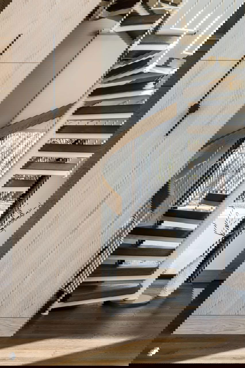 Спиральная лестница с деревянными ступенями и белой металлической конструкцией, вид на входную зону и естественный свет из окон.