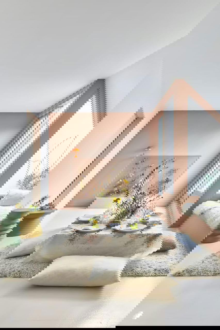 Светлая столовая зона с низким столом, покрытым скатертью в нежных тонах; на фоне пастельная стена и высокие окна, интерьер в скандинавском стиле с флористической композицией