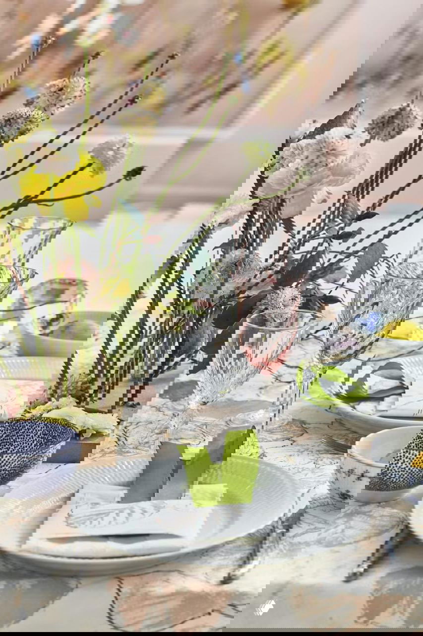 Детальный кадр сервировки: стеклянные бокалы розового оттенка, тарелки на скатерти с отпечатками листьев и живые цветы в композиции на столе